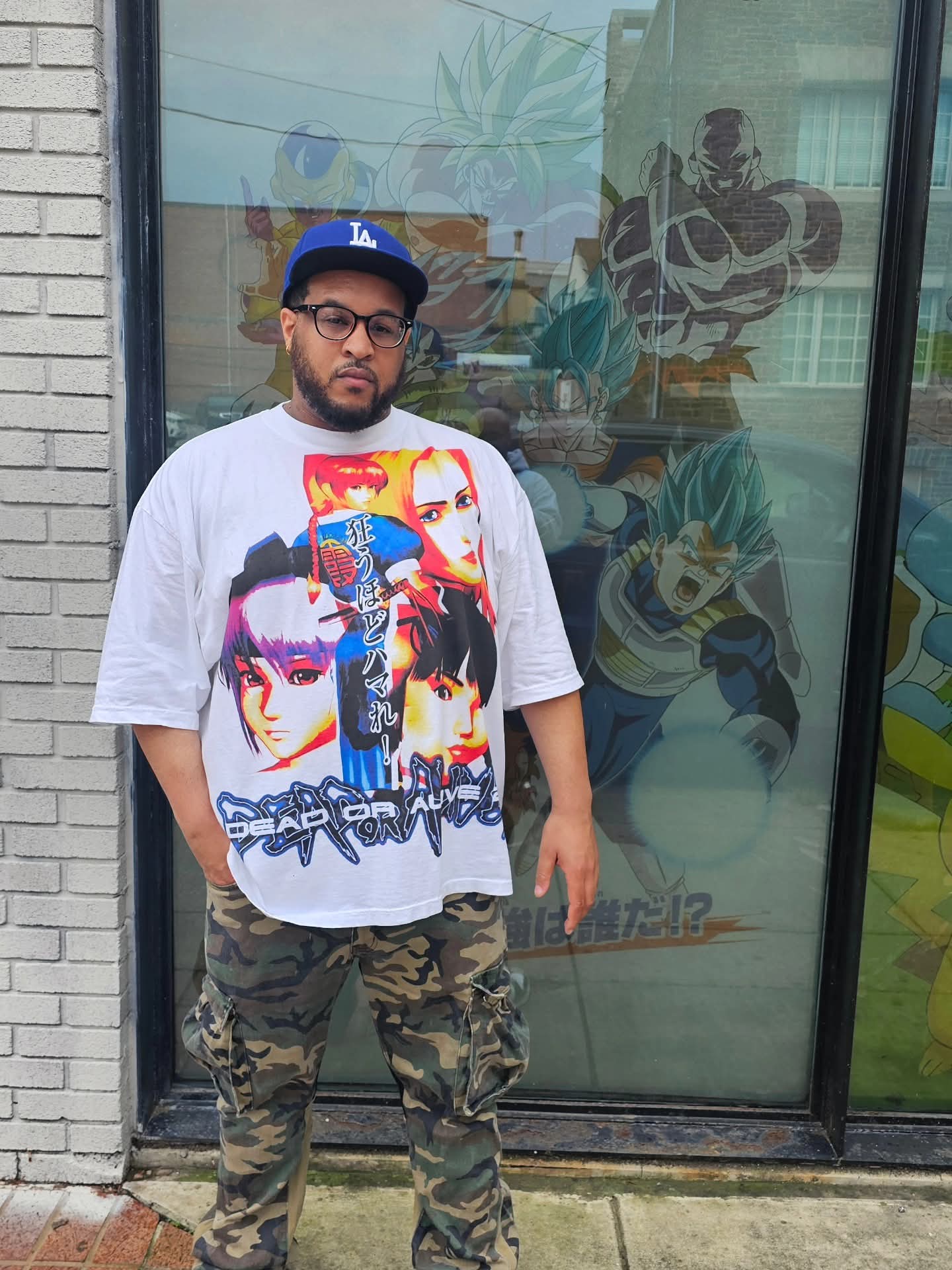 Man with glasses, beard, wearing a Los Angeles Dodgers cap, oversized graphic T-shirt, and camouflage pants, standing outside a glass door.