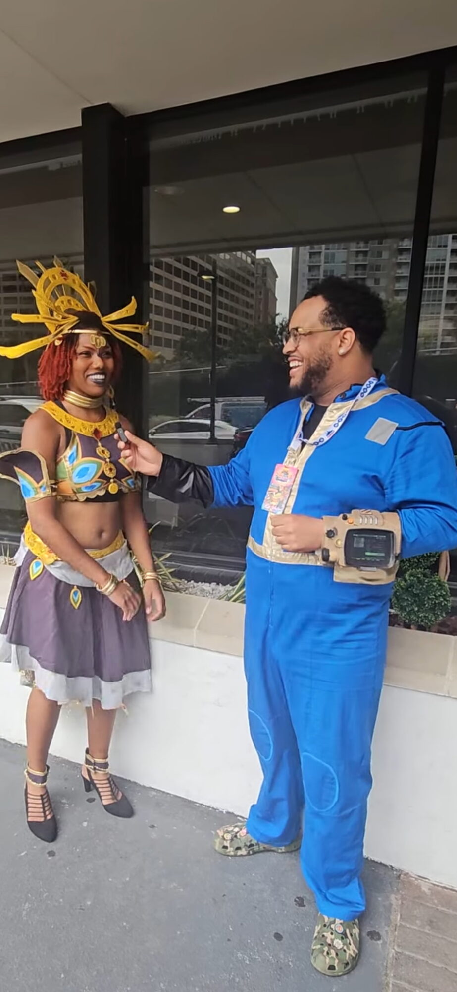 Two people in costumes stand outside a building, smiling and talking, with a cityscape in the background.