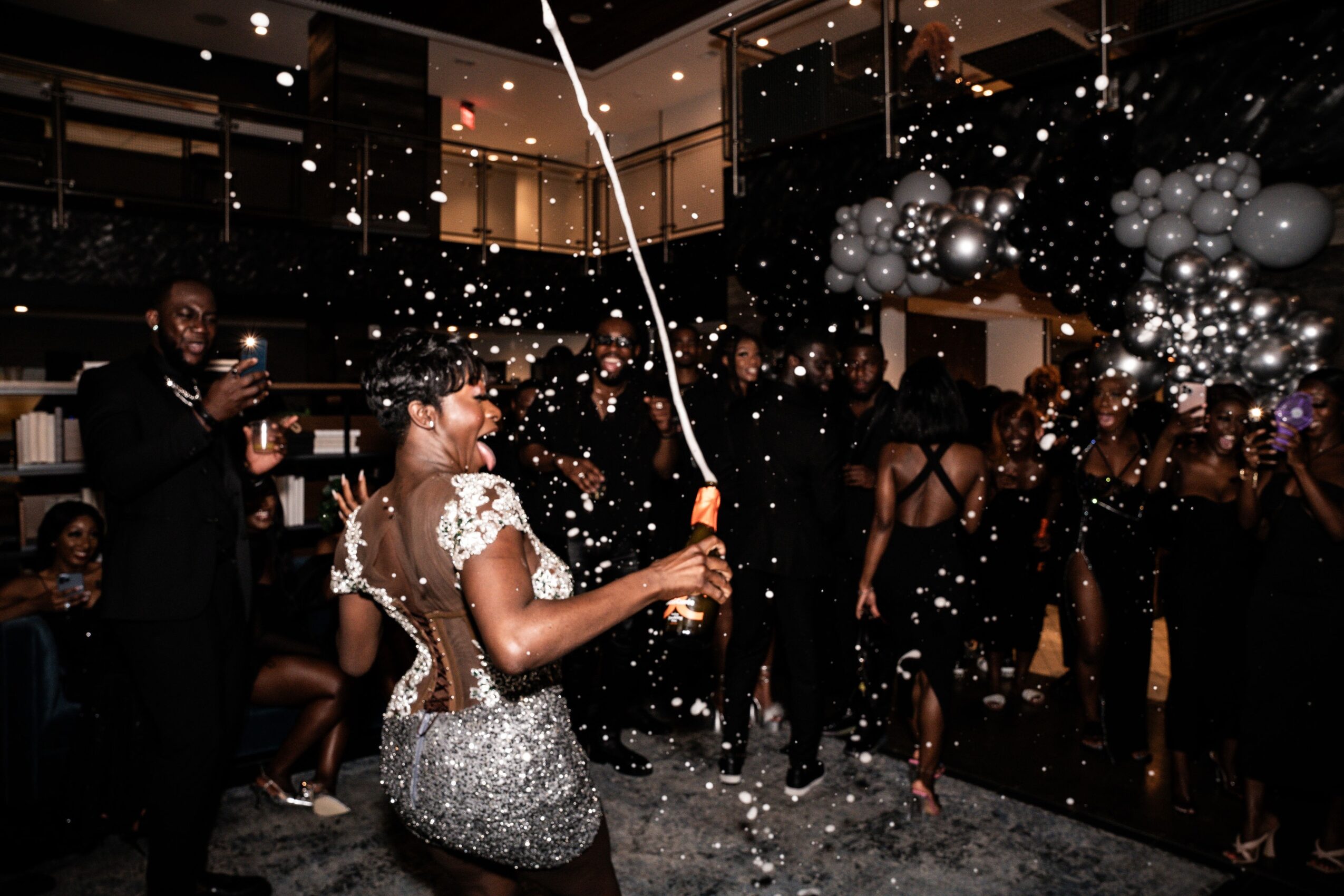 Woman holding a balloon string with confetti and balloons around, party scene with people dancing and celebrating indoors.