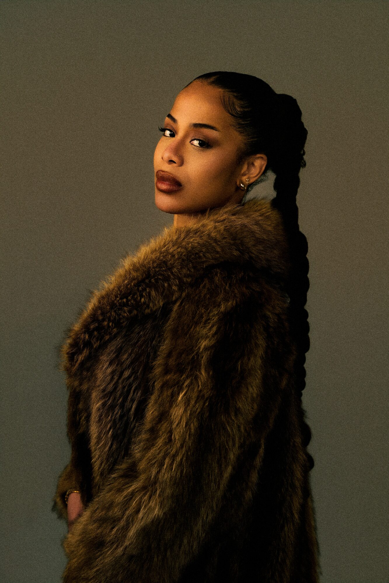 Woman with braided hair wearing a fur coat, looking at the camera against a plain background.