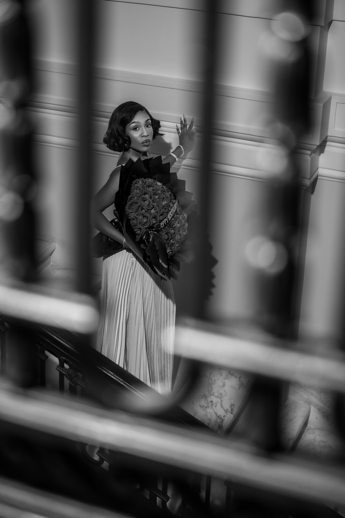 Woman with short hair in a dress holding a bag, standing behind bars, looking to the side, in black and white.