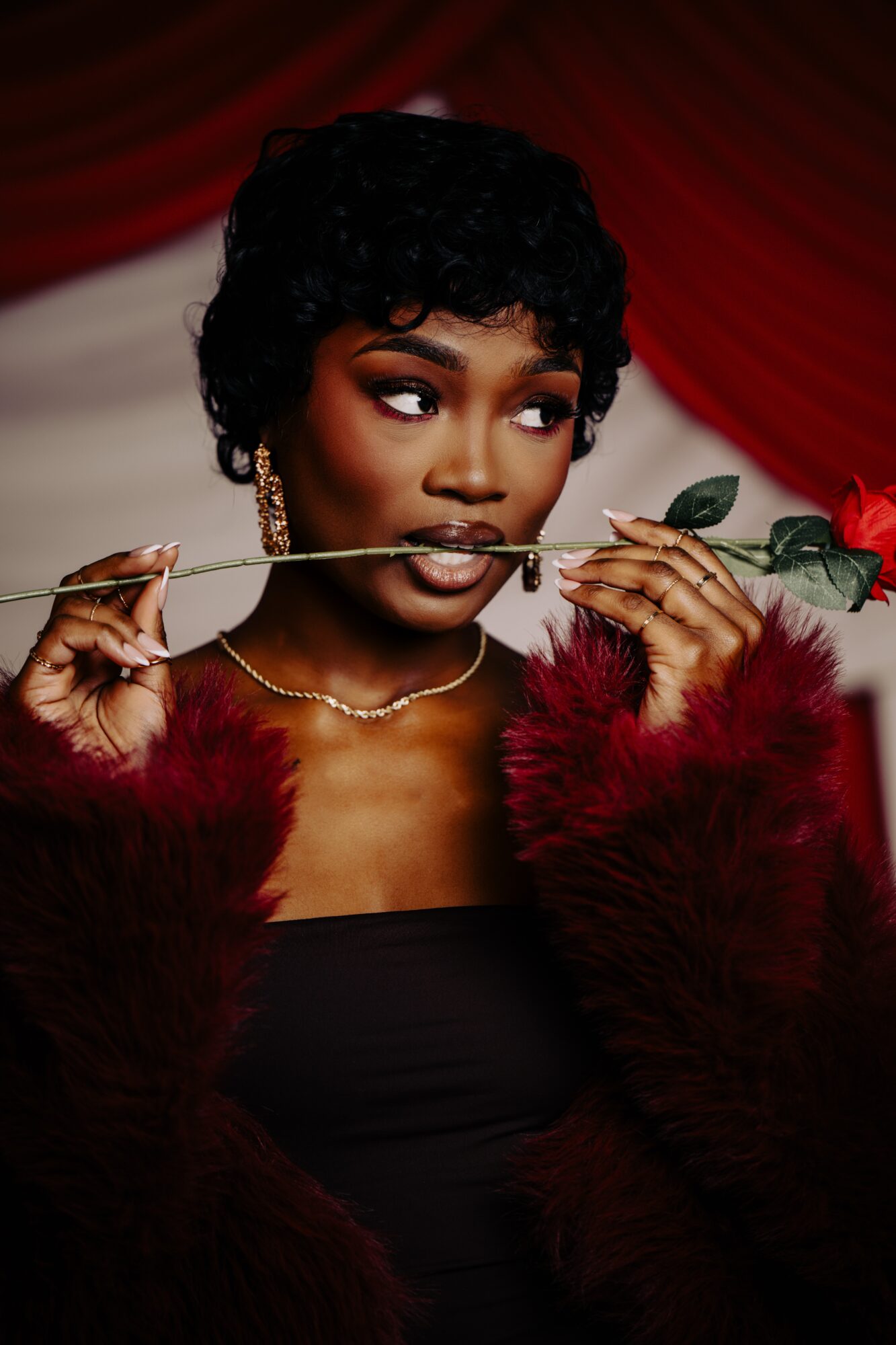 Woman with short curly hair holding a rose in her mouth, wearing a red fur coat and jewelry, against a red curtain background.