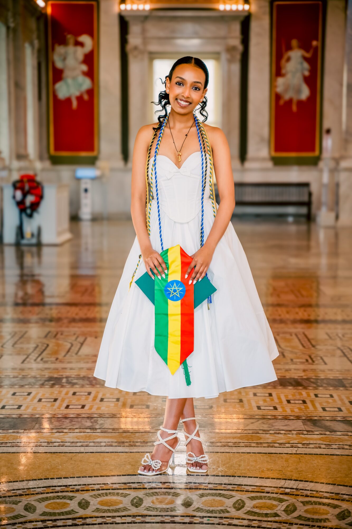 Young woman in white dress holding a Brazilian flag, standing in a grand hall with red banners and marble columns.