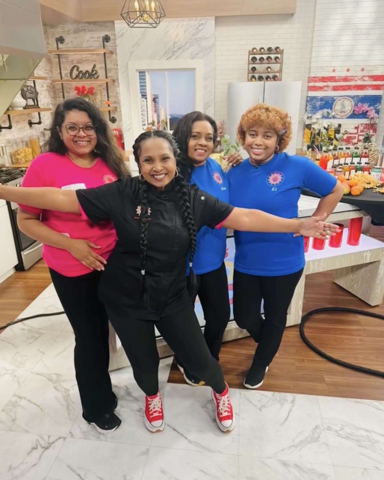 Four smiling women standing together indoors, arms around each other, in a decorated room with shelves and tables.