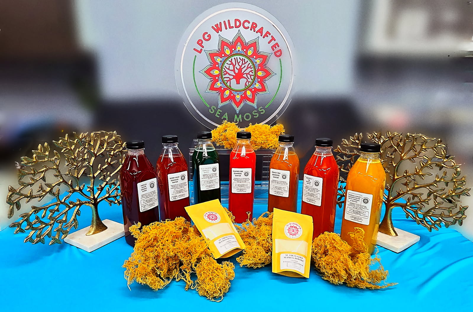 Display of colorful bottles, yellow flowers, and decorative trees with a logo in the background, on a blue tablecloth.