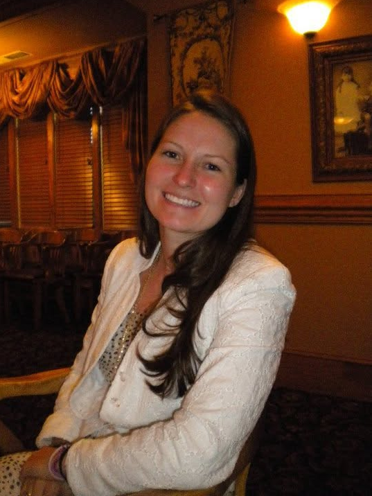 Smiling woman with long dark hair in a white blazer in a warmly lit room with wooden blinds and framed artwork.