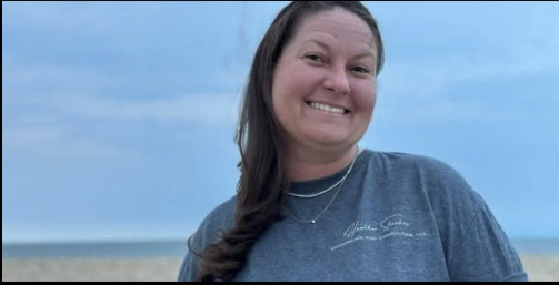 Smiling woman with long dark hair outdoors against a blue sky, wearing a blue shirt and necklace.
