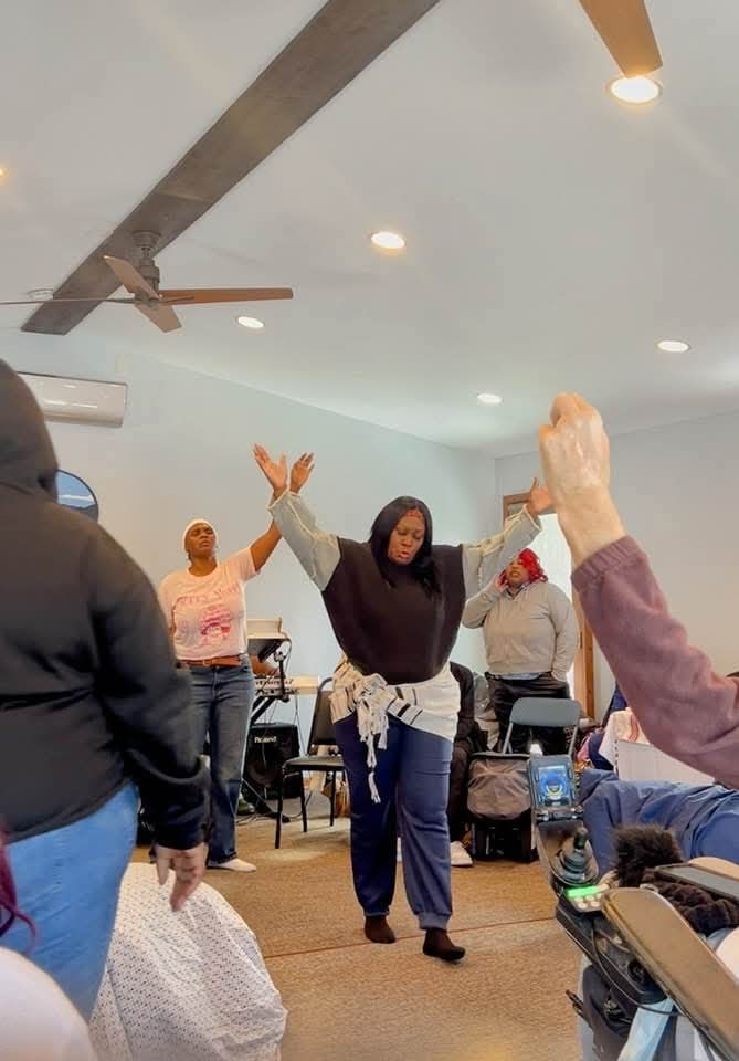 Woman with arms raised in a room with several people, ceiling fan, and chairs, celebrating or dancing.