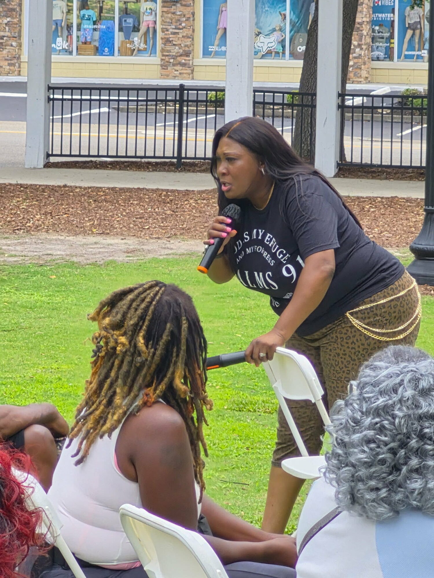 Woman speaking into a microphone outdoors, with people seated nearby and a grassy area behind her.