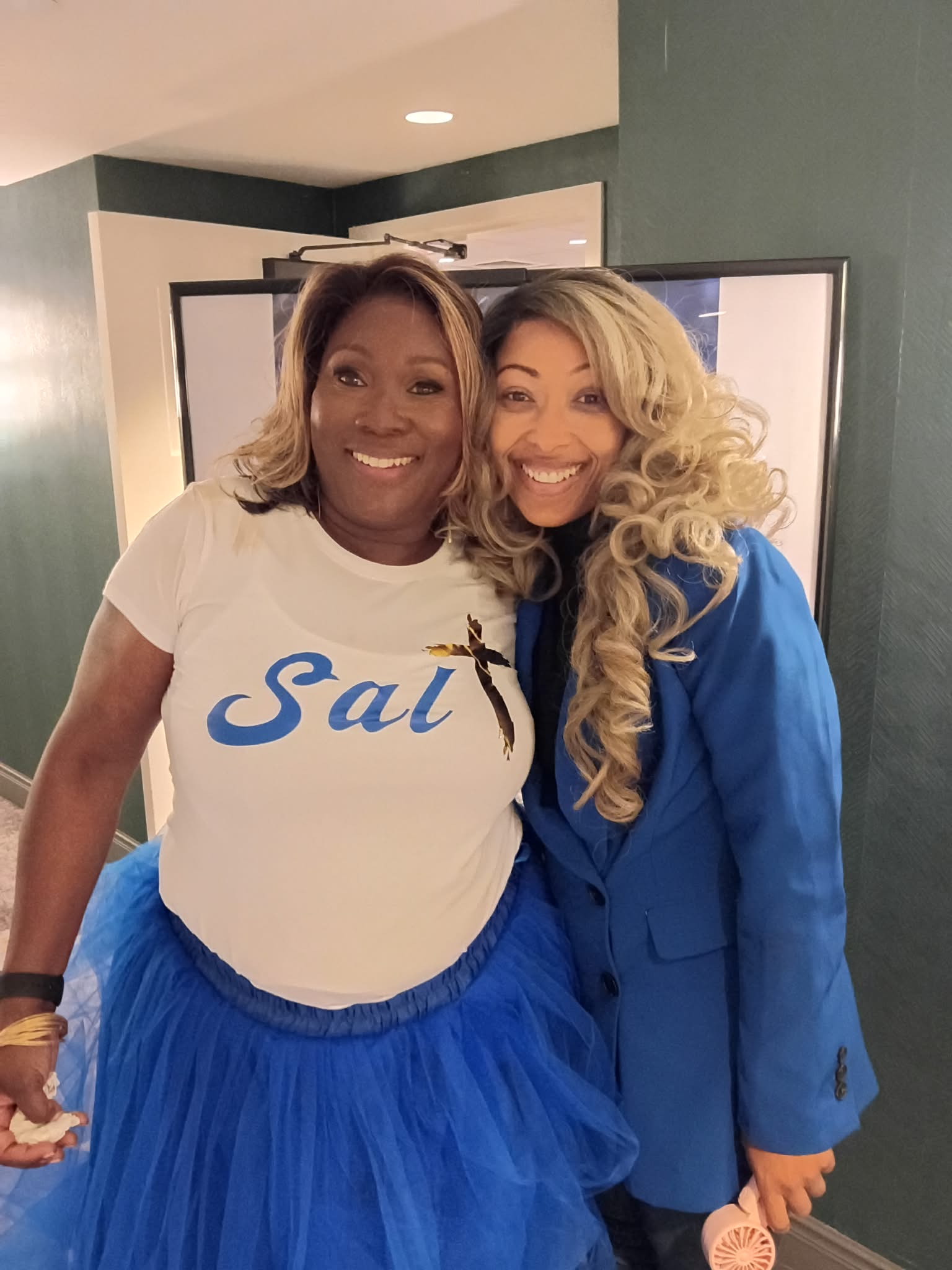 Two women smiling, one wearing a white t-shirt with blue text and a blue skirt, the other in a blue blazer and dress, standing close together.