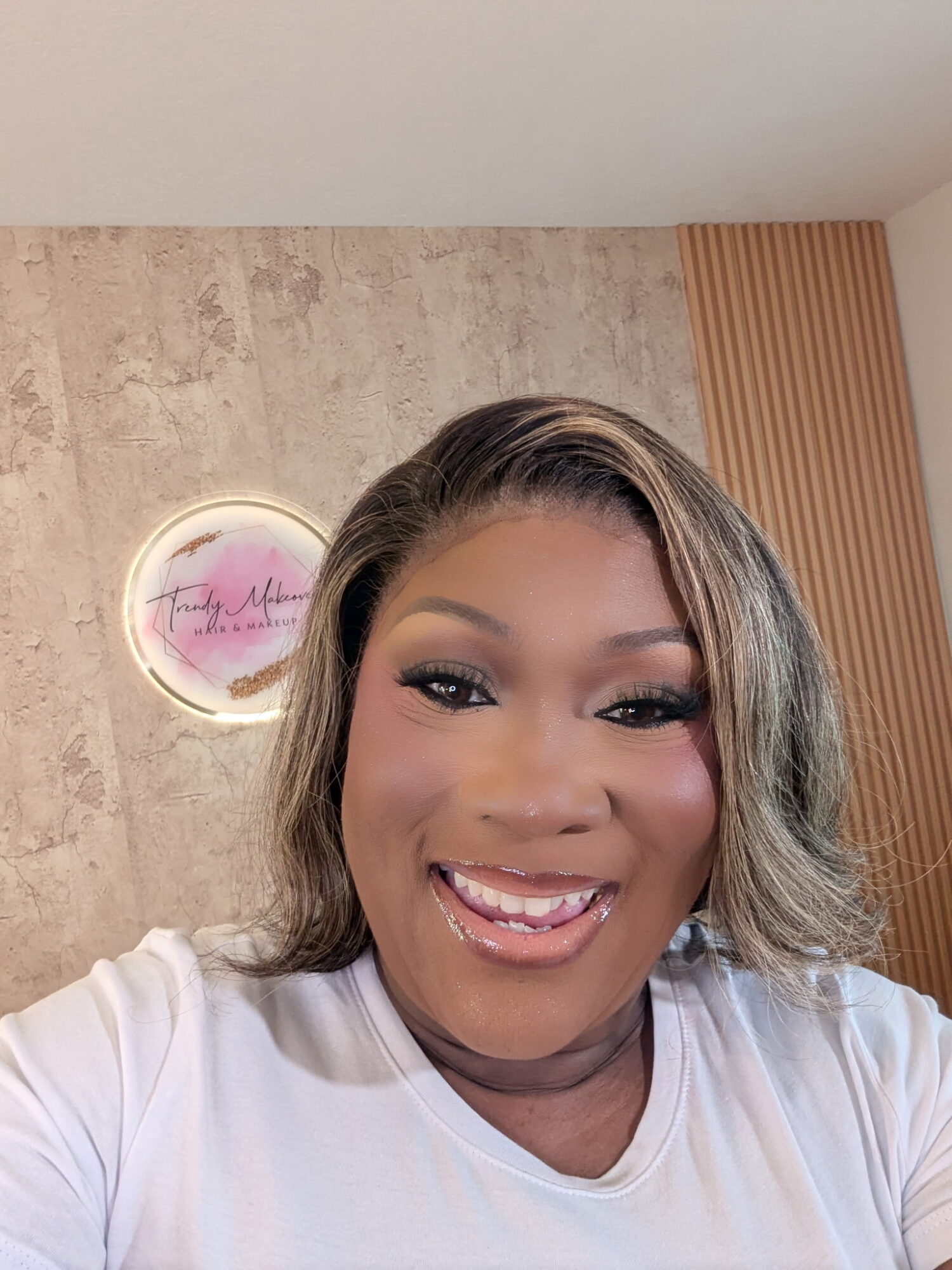 Smiling woman with makeup and wavy hair taking a selfie indoors, background includes a textured wall and a neon sign.