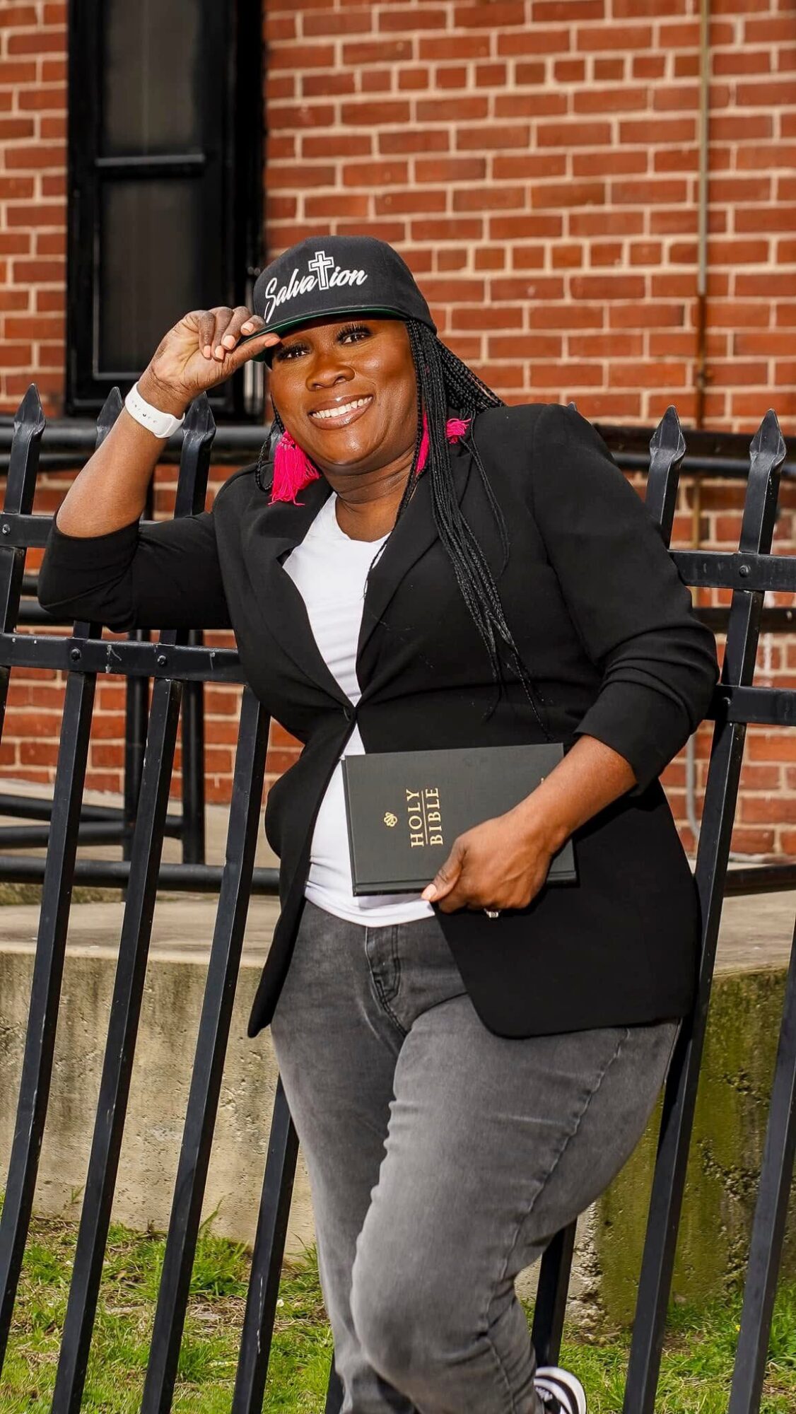 Woman smiling, wearing a black cap, black blazer, white shirt, holding a book, standing outdoors near a brick building.