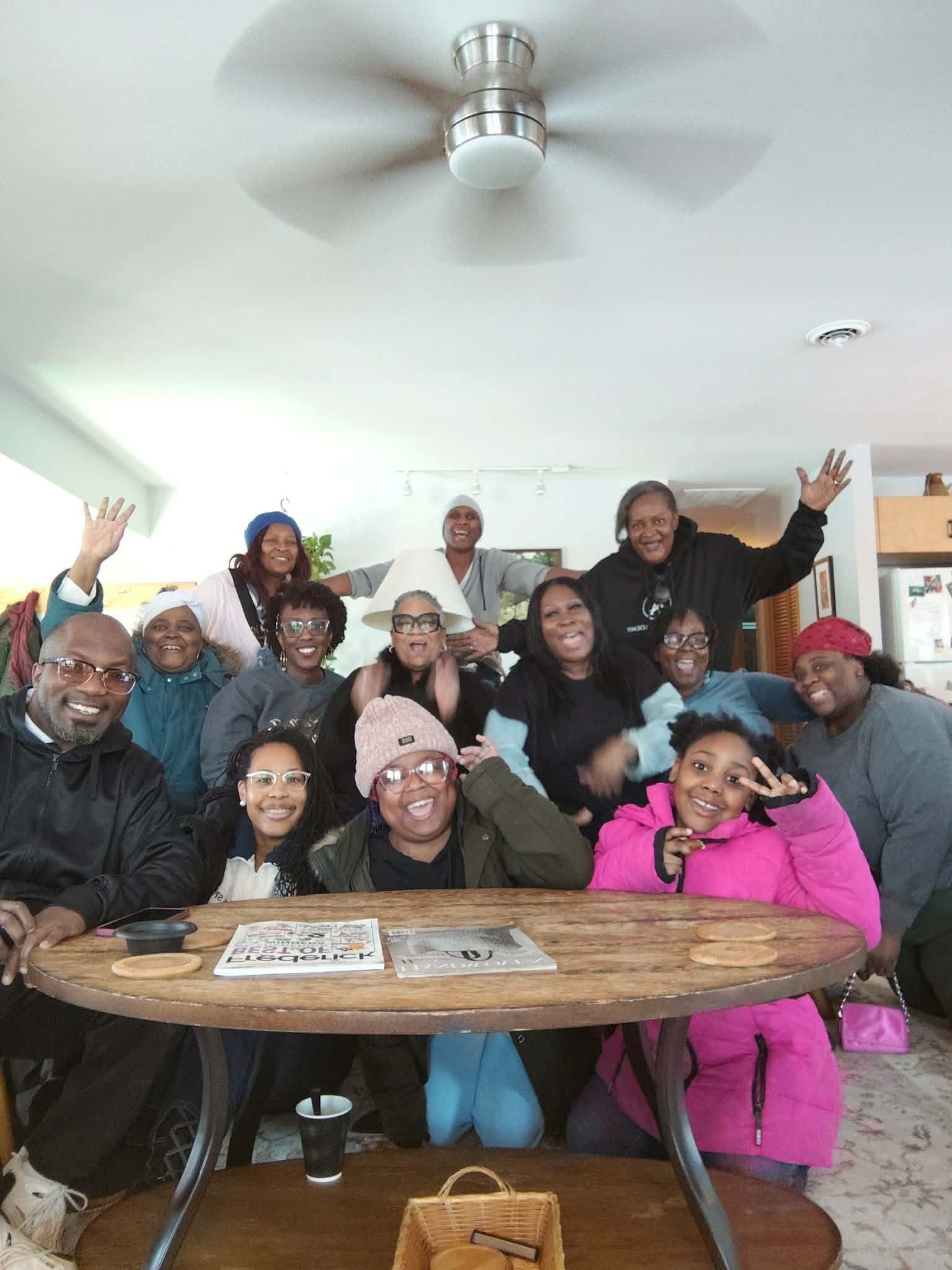 Group of people smiling and waving around a wooden table in a bright room.