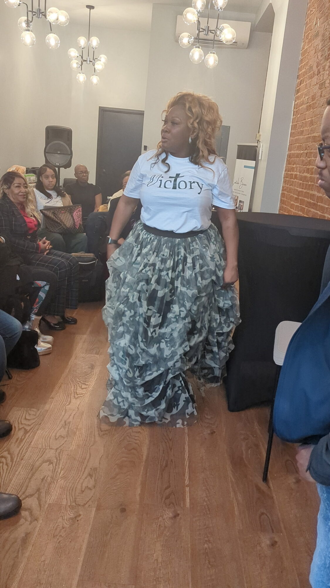 Woman in a white t-shirt and patterned skirt walking in a room with seated audience, chandeliers overhead.