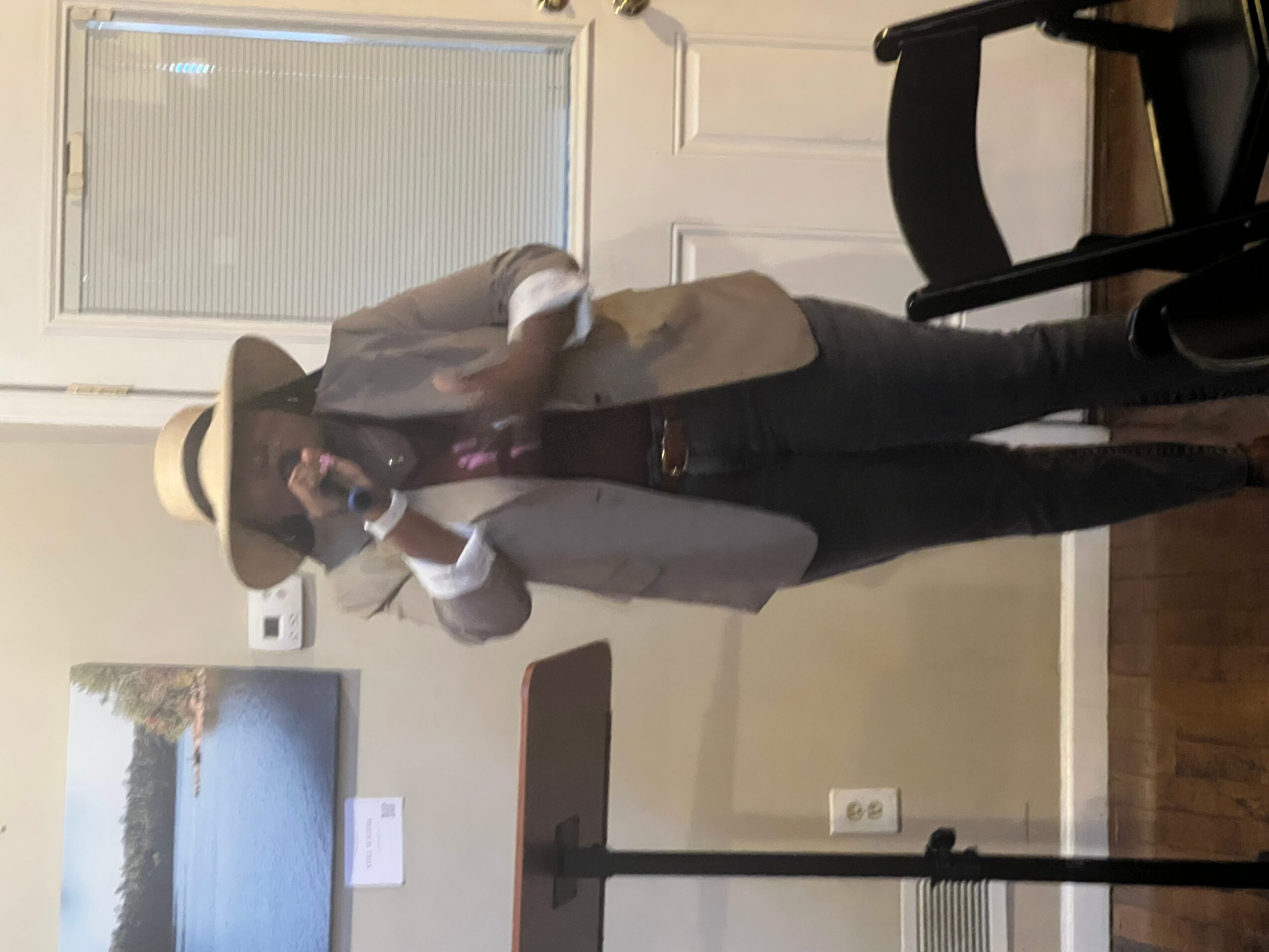 Person wearing a hat, white shirt, and vest, standing indoors near a window and a table.