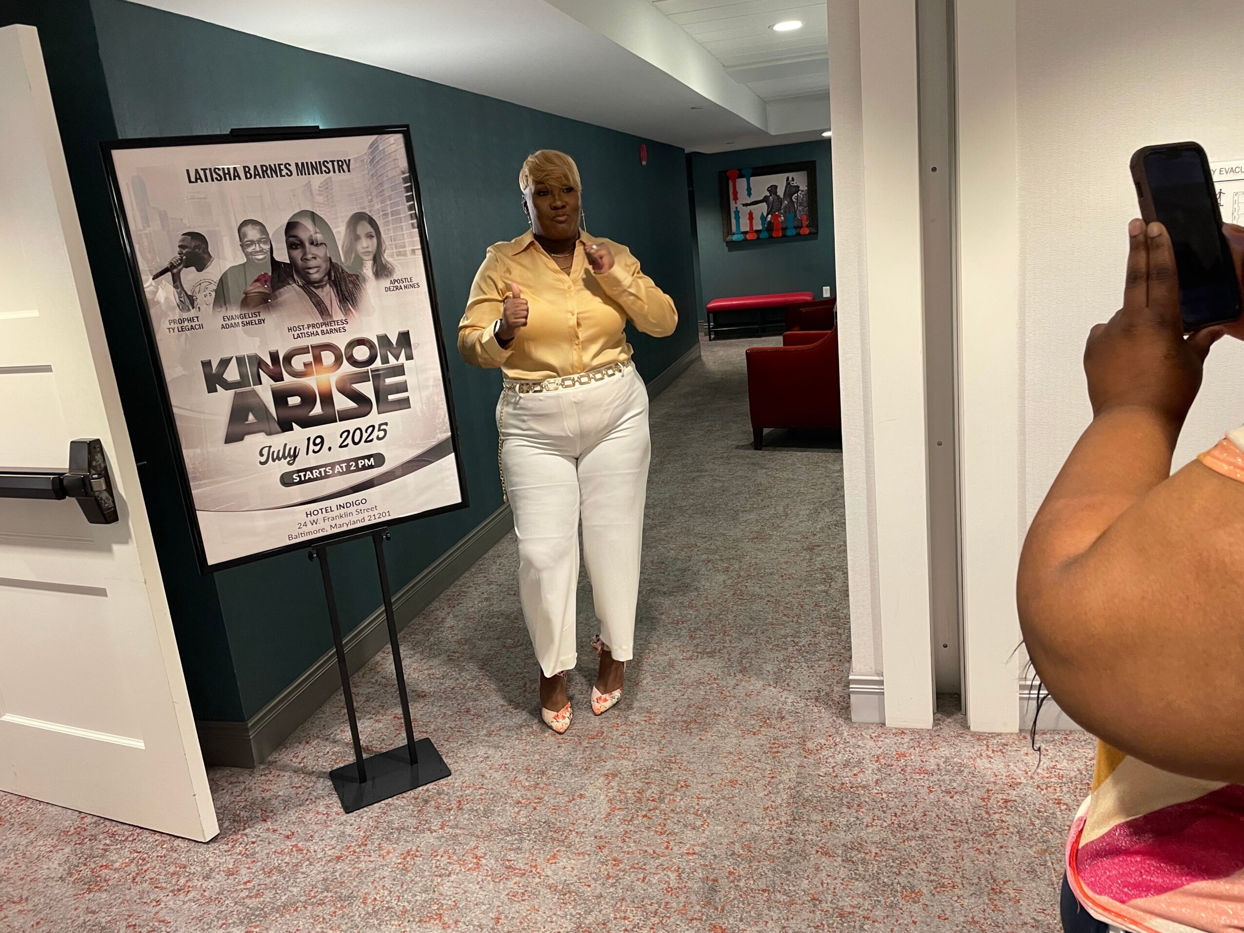 Woman standing in hallway, person taking photo with phone, poster on wall, carpeted floor, seating area in background.