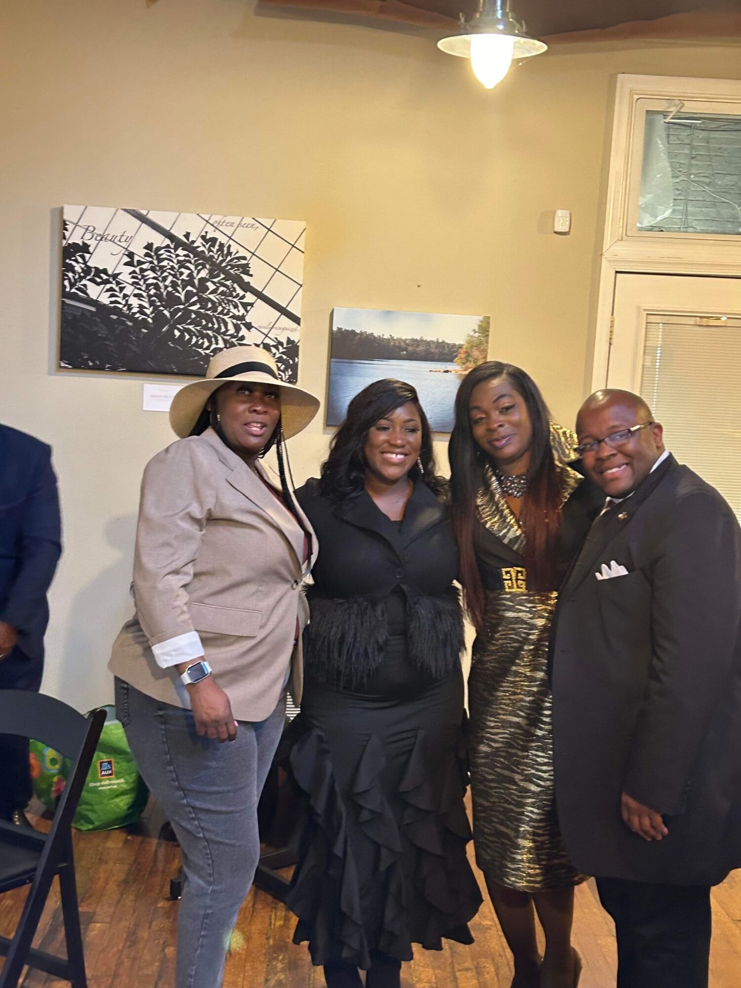 Four people standing indoors, smiling, with artwork on the wall behind them.