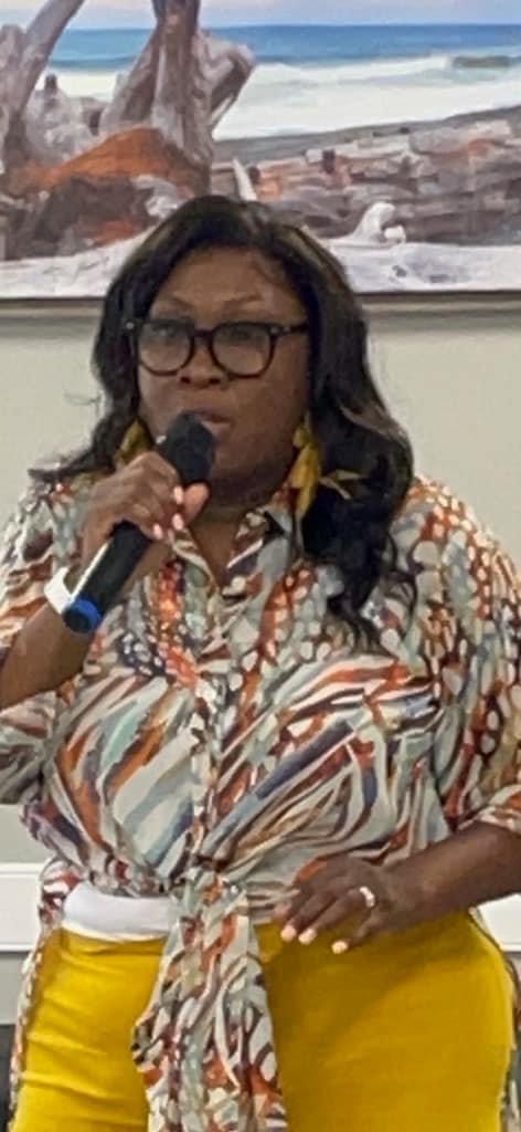Woman with glasses speaking into microphone, wearing patterned blouse and yellow pants, standing indoors with beach scene on screen behind her.