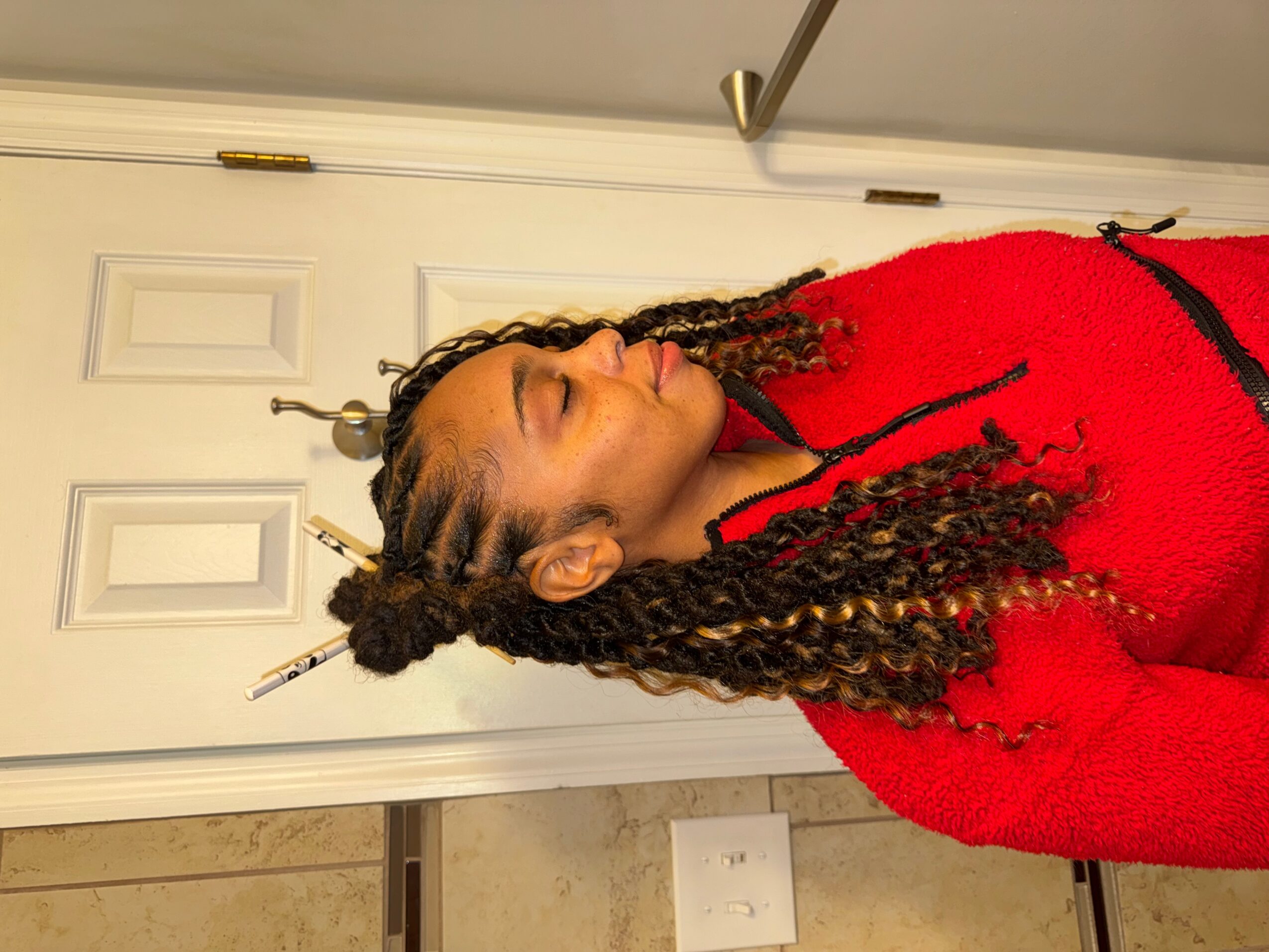 Woman with braided hair and a red sweater, standing sideways in front of a white door, eyes closed.