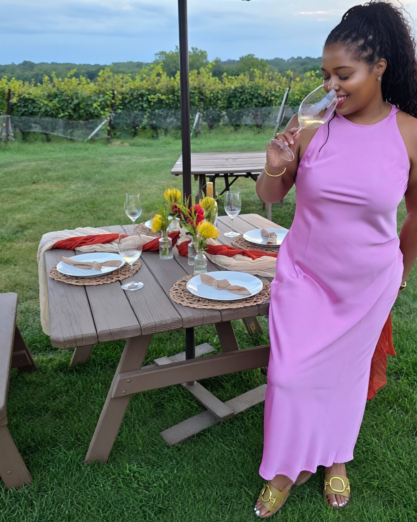 Woman in pink dress holding a glass, standing near a table set for outdoor dining with flowers and plates.