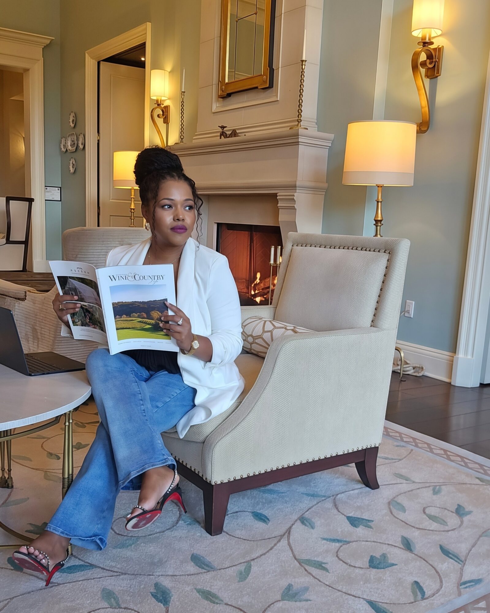 Woman sitting in a cream armchair reading a magazine in a warmly lit living room with fireplace and wall sconces.