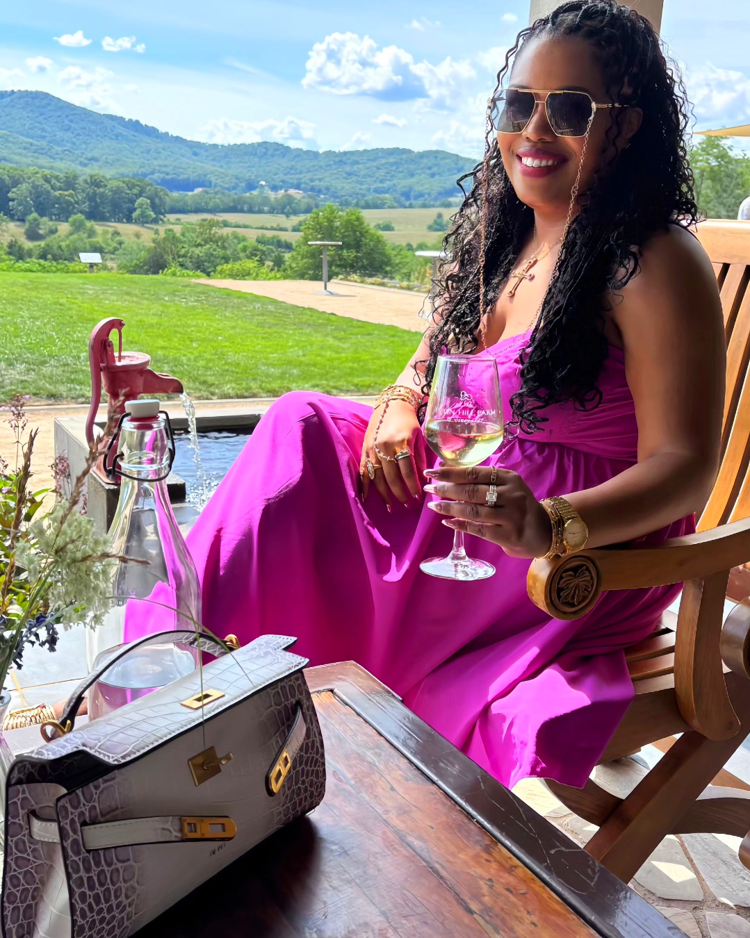 Woman in pink dress with sunglasses sitting outdoors holding a glass, with a scenic landscape background.