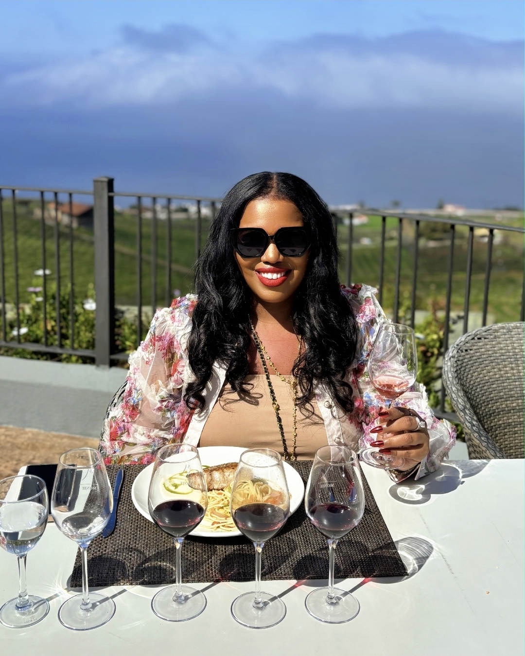 Woman with long dark hair, sunglasses, and red lipstick, smiling at a table with wine glasses outdoors.