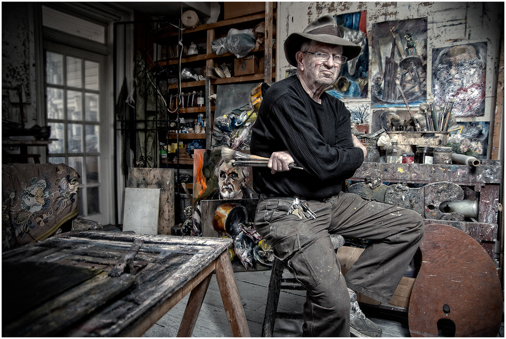 An artist wearing a hat sits in a cluttered studio with paintings and art supplies around him.