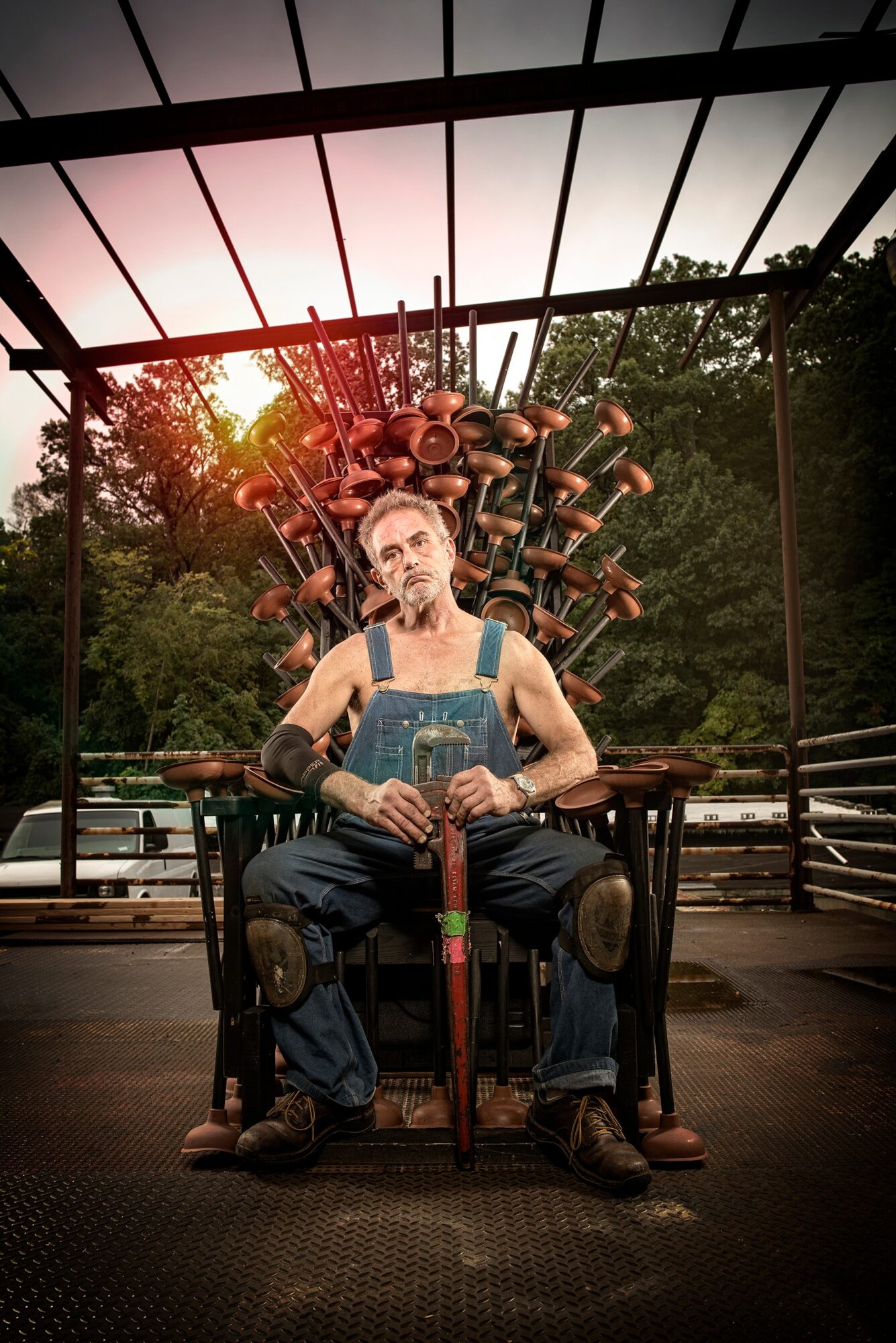 Man sitting on a throne-like chair with a large sculpture of musical instruments behind him outdoors, sunset in the background.