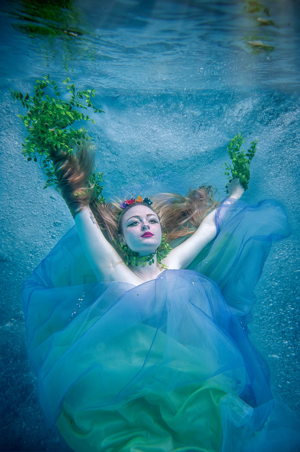 Person in a flowing blue dress underwater with arms raised, surrounded by bubbles and green plants, face visible with makeup.