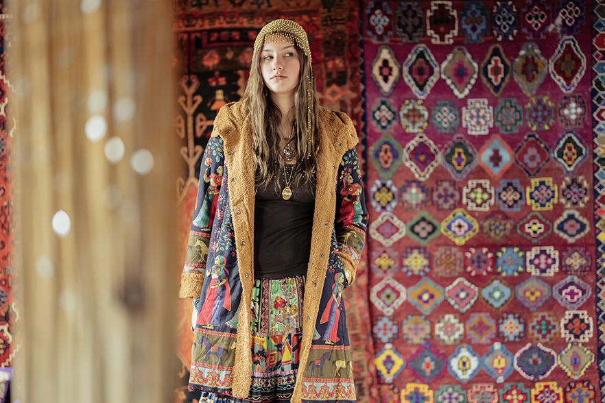 Young woman with long hair wearing a colorful, patterned jacket and headband, standing in front of a vibrant, patterned rug.