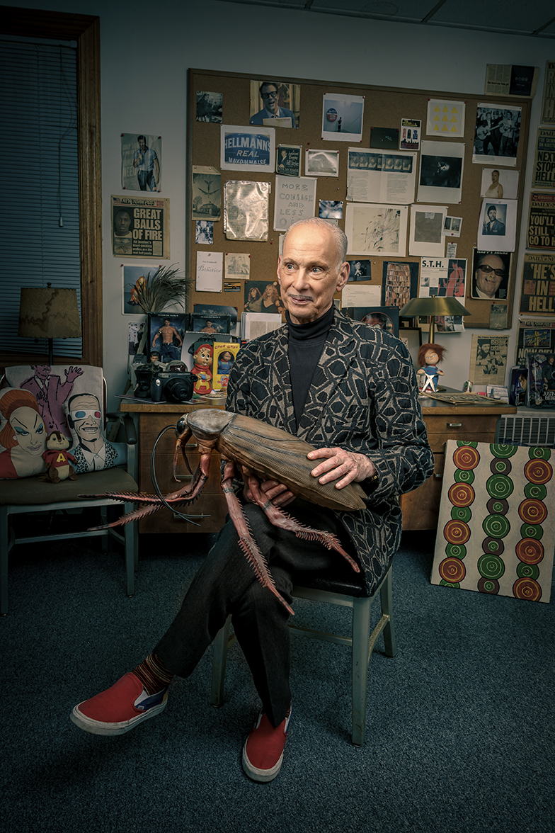Man sitting on a chair holding a wooden object in a room with a collage of photos and artwork on the wall behind him.
