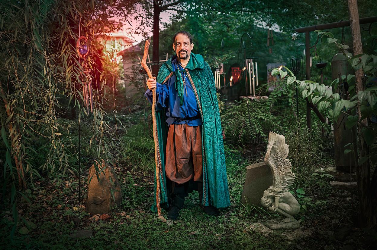 Person dressed as a wizard holding a staff in a garden with trees and a stone sculpture, sunlight filtering through trees.