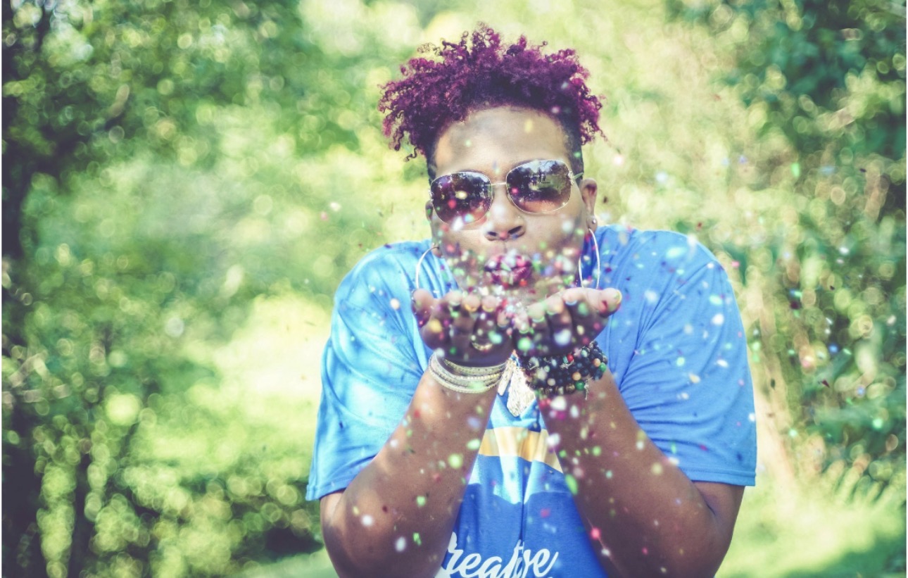 Person with curly hair wearing sunglasses and a blue shirt blowing colorful confetti outdoors.