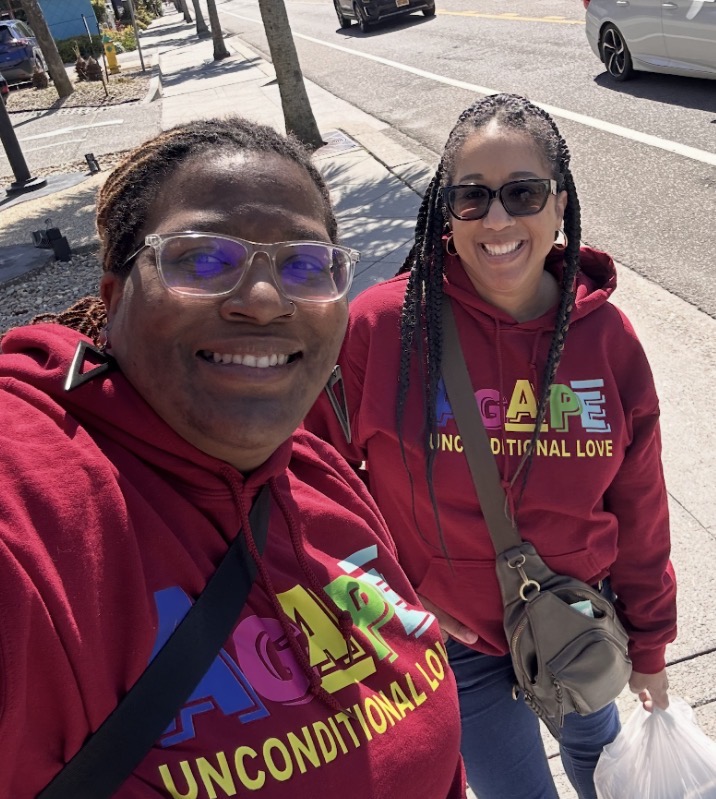 Two smiling women wearing matching maroon hoodies with colorful text, standing on a sidewalk with trees and cars in the background.