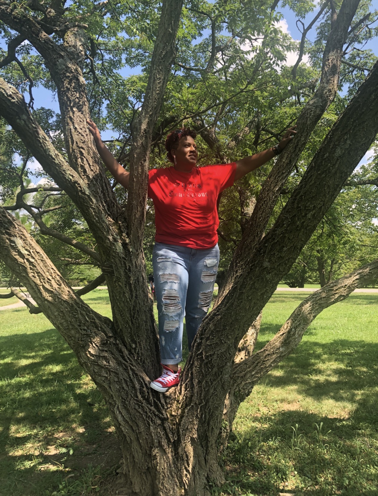 Person standing on a tree branch with arms outstretched, wearing a red shirt and ripped jeans, in a park with green grass and trees.