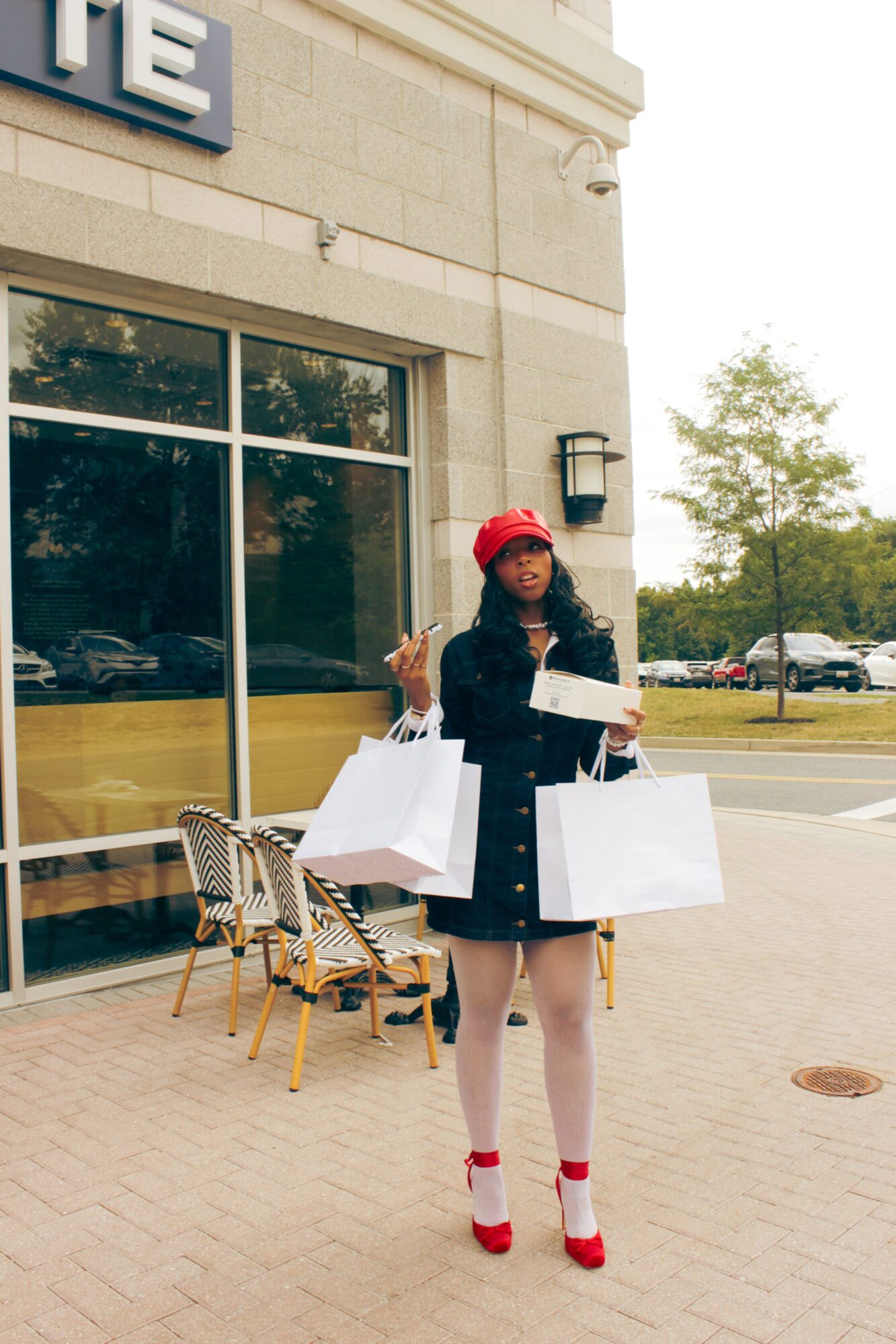 Woman in red hat and black coat holding shopping bags outside store, with chairs and large window behind her.