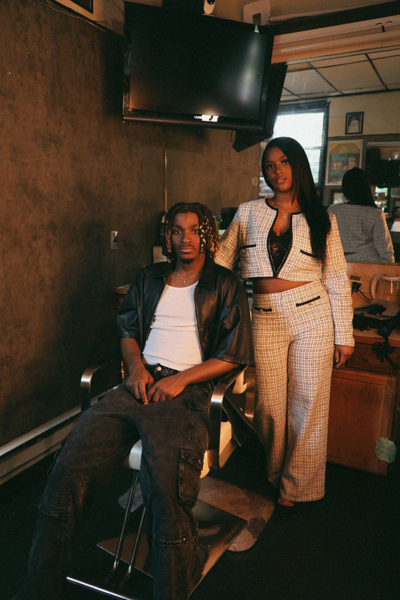 Two women pose indoors, one seated and one standing, with a TV and window in the background.