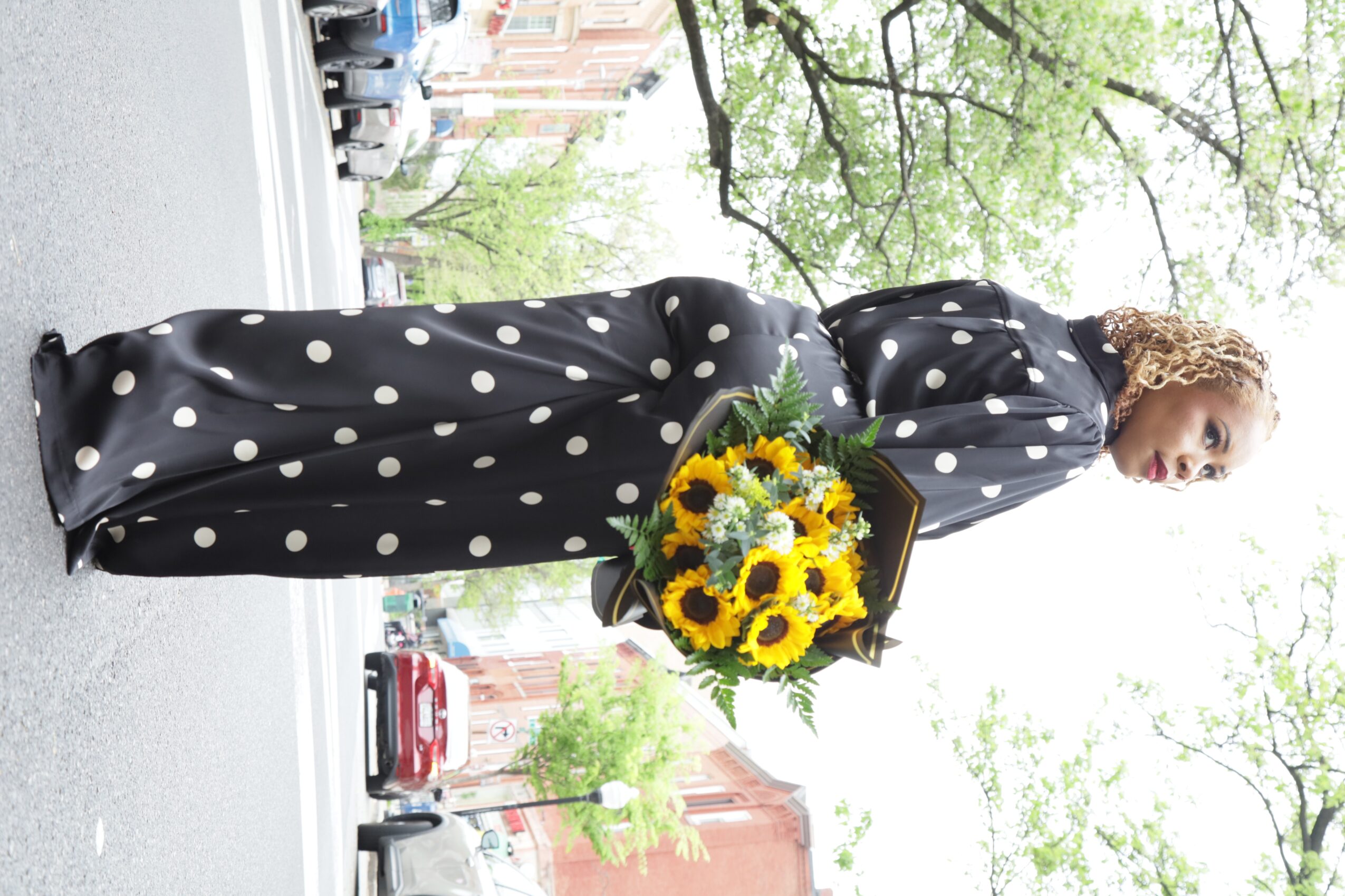 Woman holding a sunflower bouquet on a city street, trees and cars in background, wearing a black polka dot dress.
