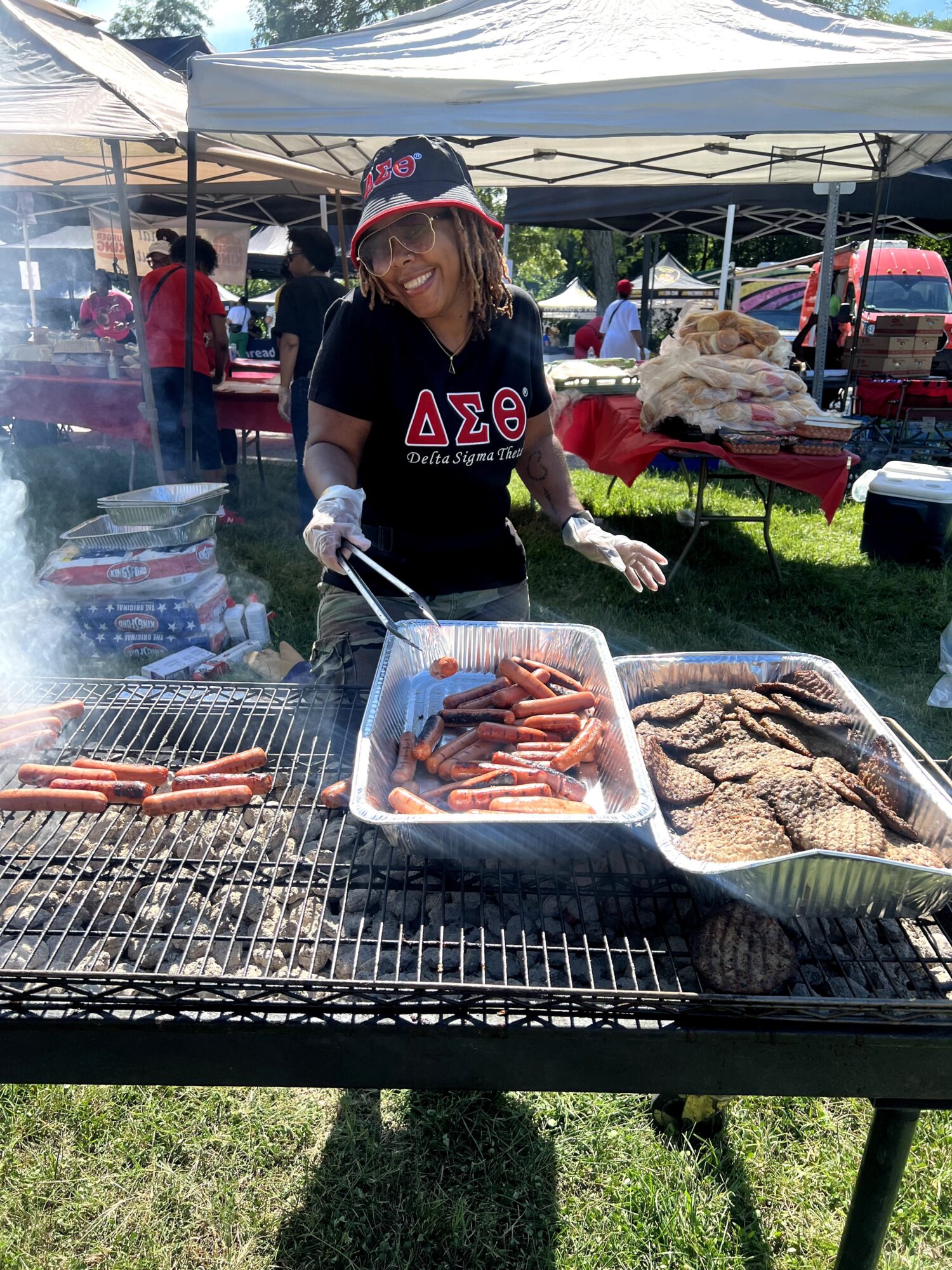 Person grilling hot dogs and sausages at an outdoor event under a canopy, with other tents and people in background.