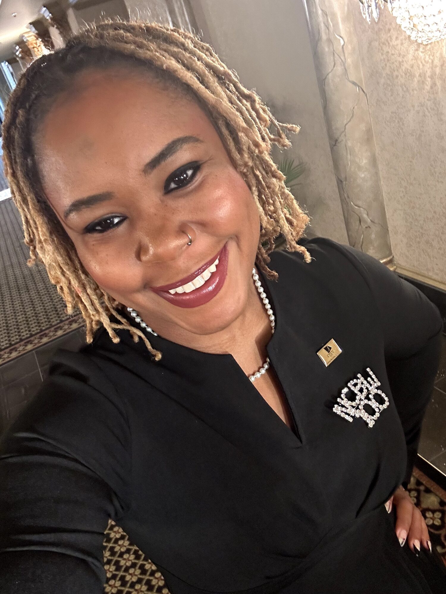 Smiling woman with blonde dreadlocks wearing a black uniform and pearl necklace.