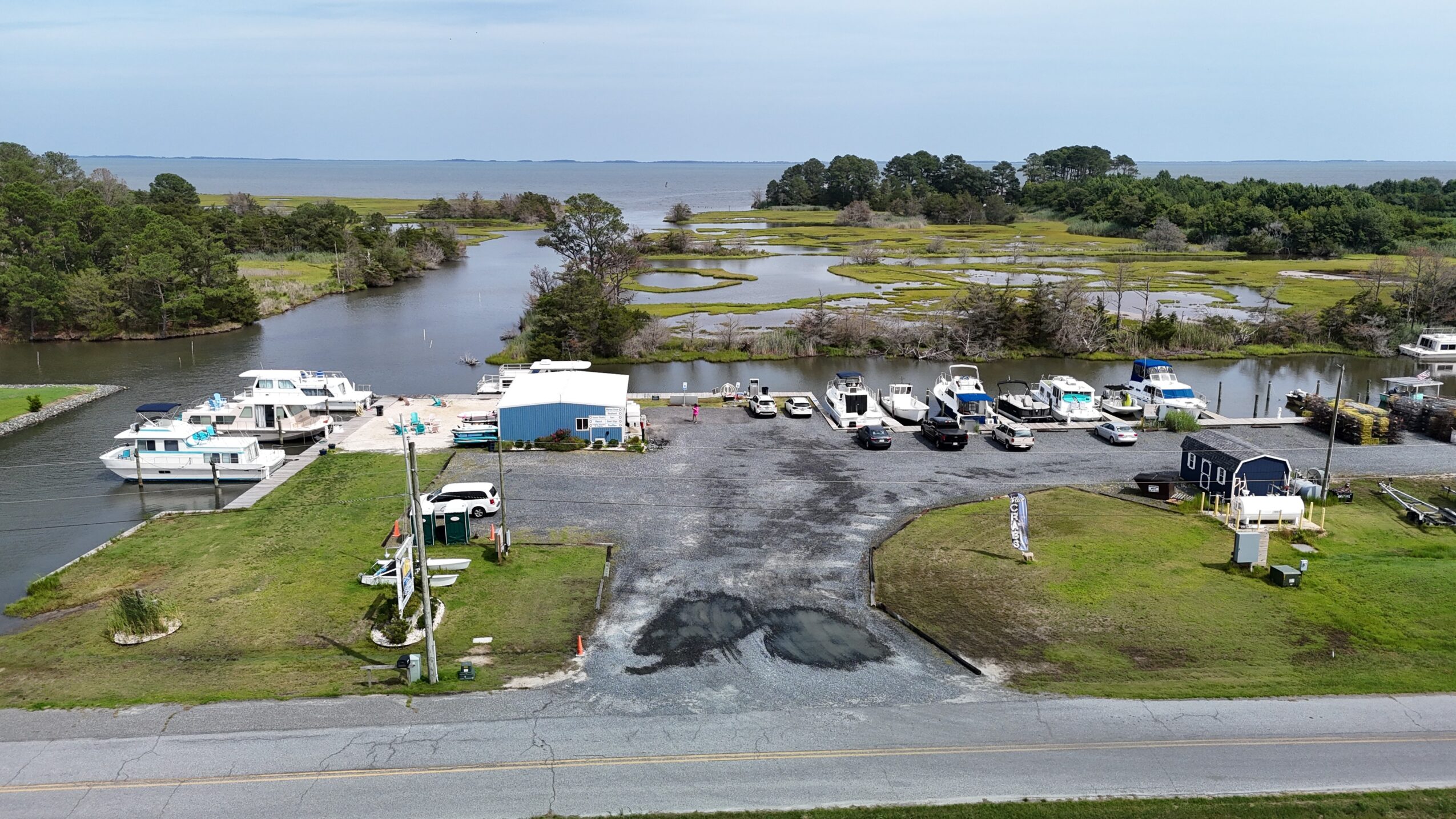 Parking lot with boats, vehicles, and buildings near water, with marshes and trees in the background.