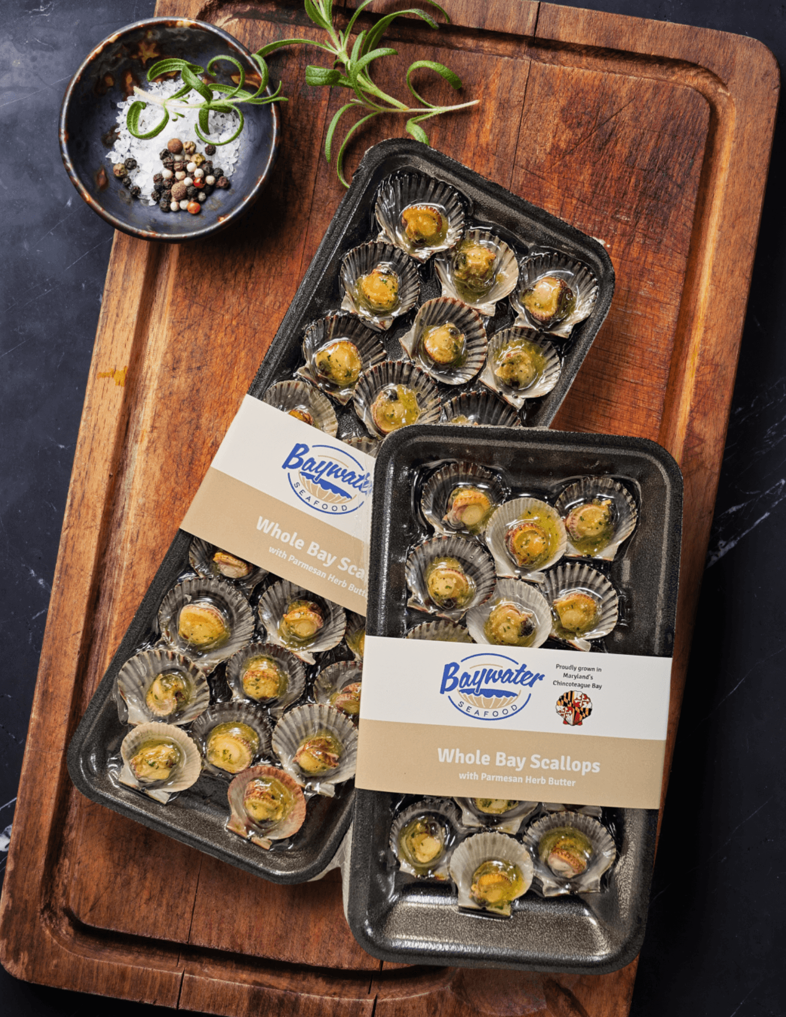 Two trays of cooked scallops on a wooden cutting board, with a bowl of sauce and a plant nearby.