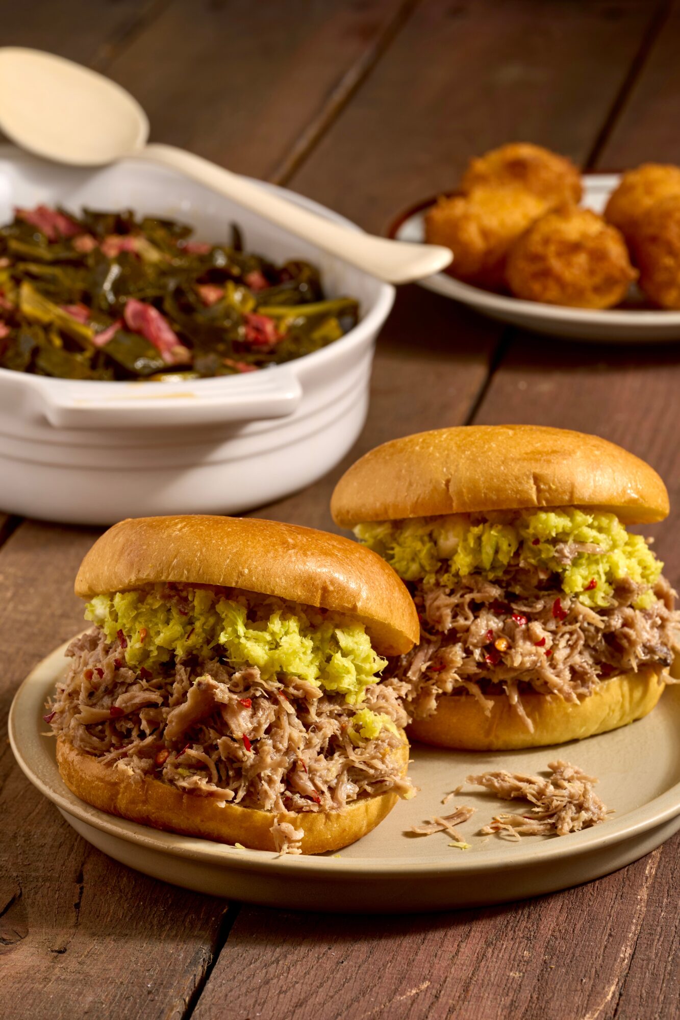 Two sandwiches with shredded meat and pickles on a plate, with fried food in the background, on a wooden table.