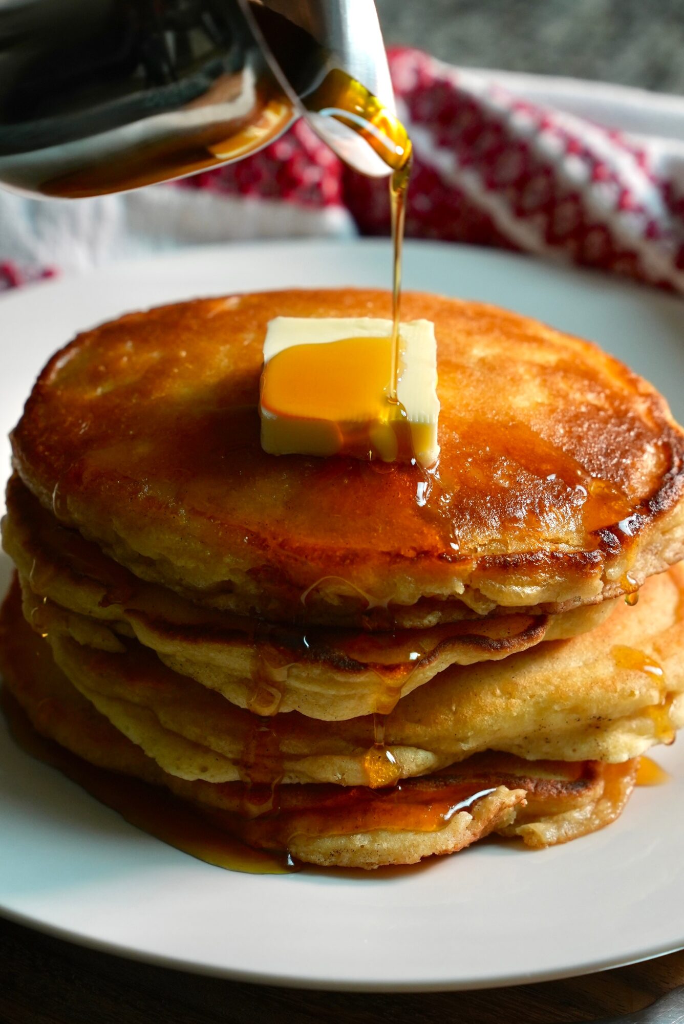 Stack of pancakes with butter, syrup being poured from a bottle, on a white plate with a red and white cloth in background.