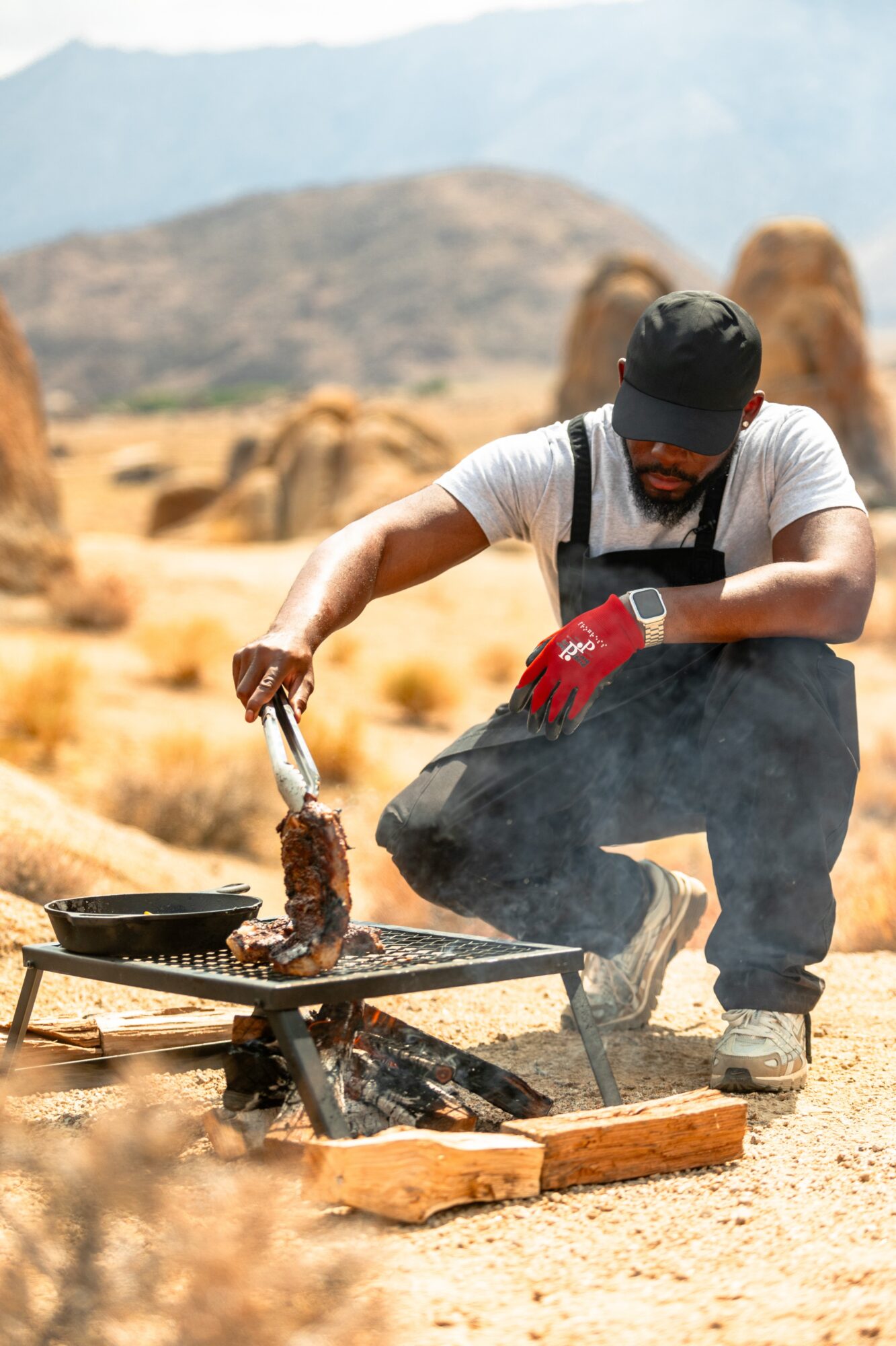 Person grilling meat outdoors in a desert landscape with rocks and mountains in the background.