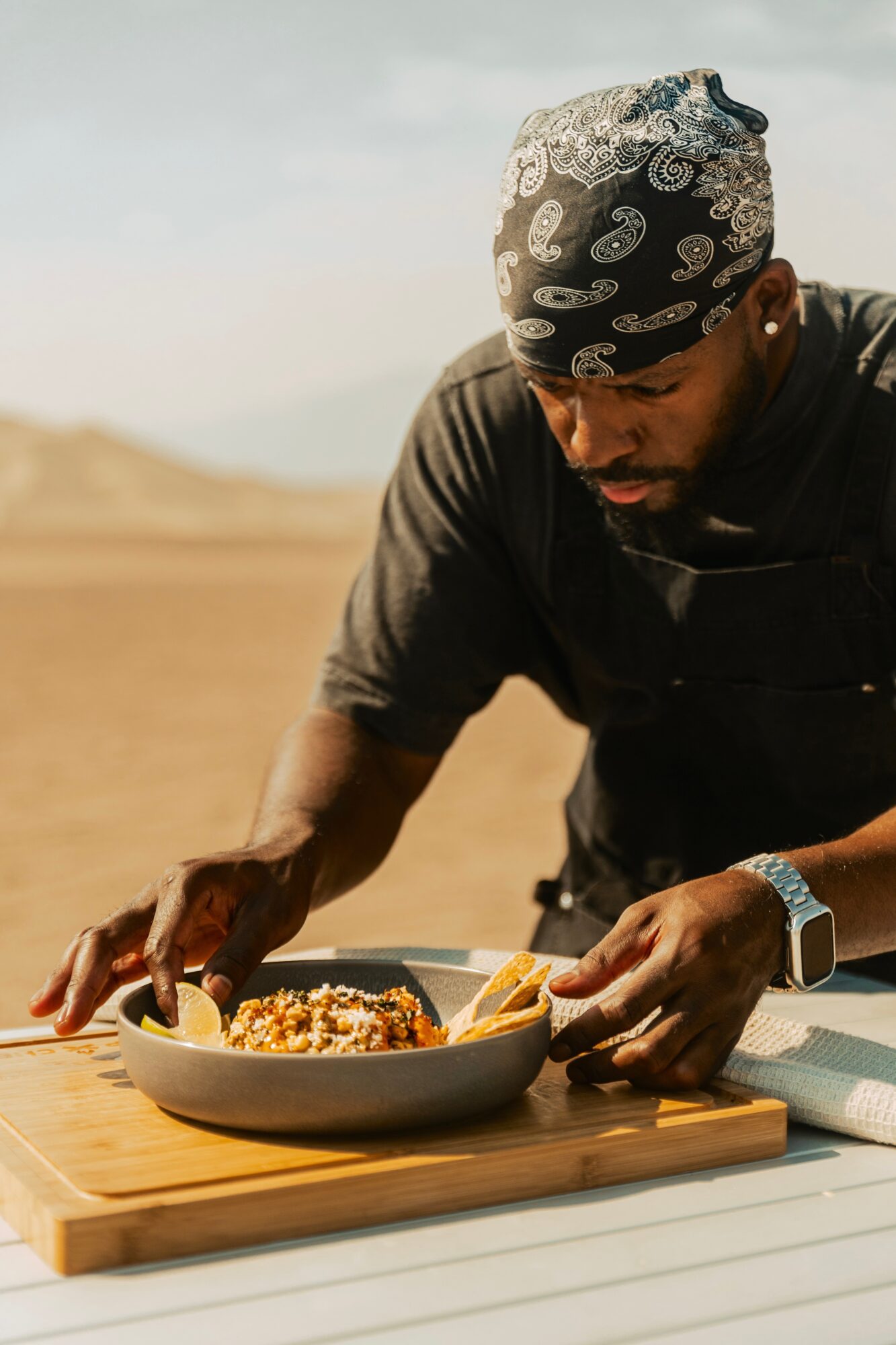 Person wearing a bandana and black shirt preparing food outdoors, with a bowl on a wooden surface.
