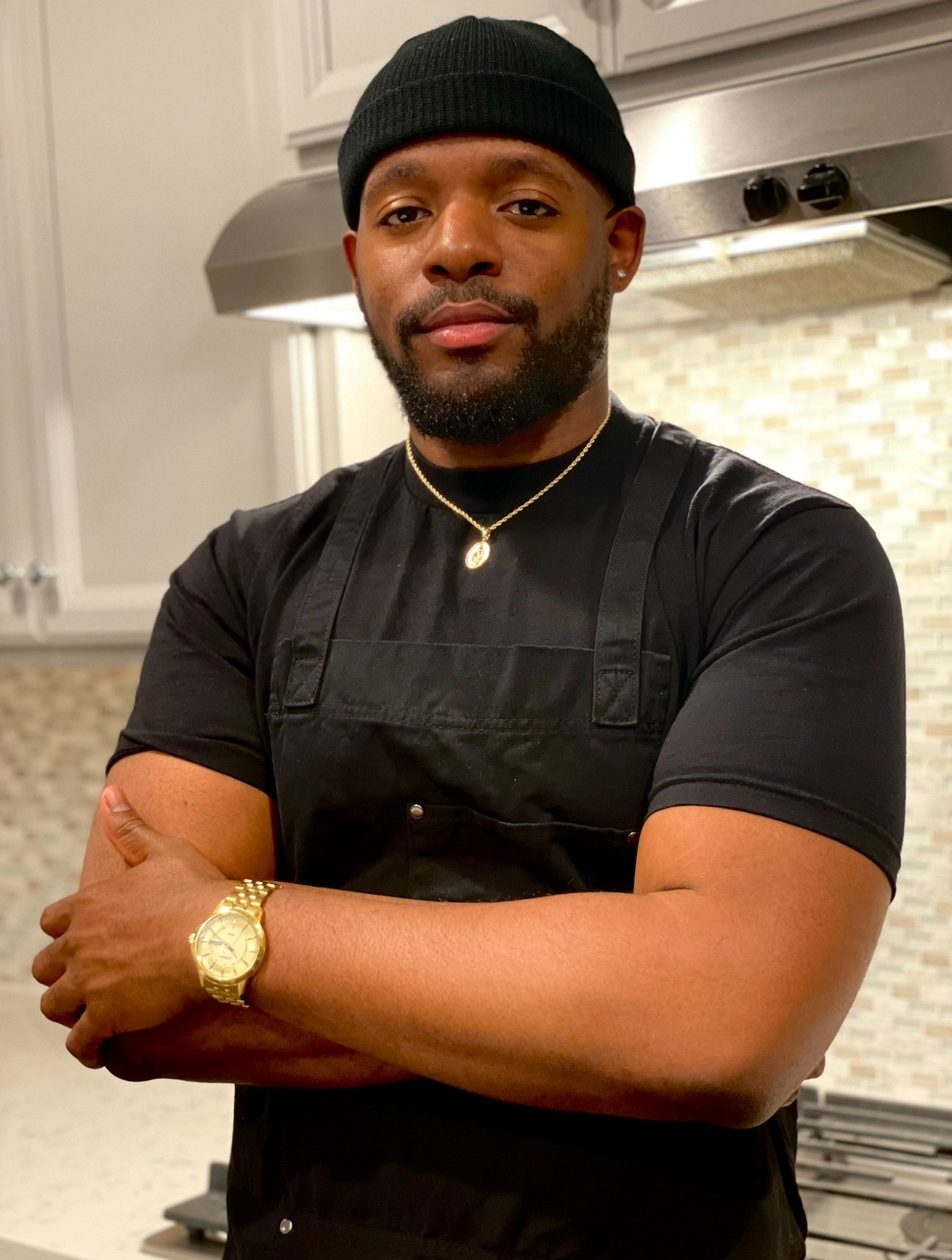 Man with a beard wearing a black beanie, black shirt, and apron, standing in a kitchen with arms crossed.