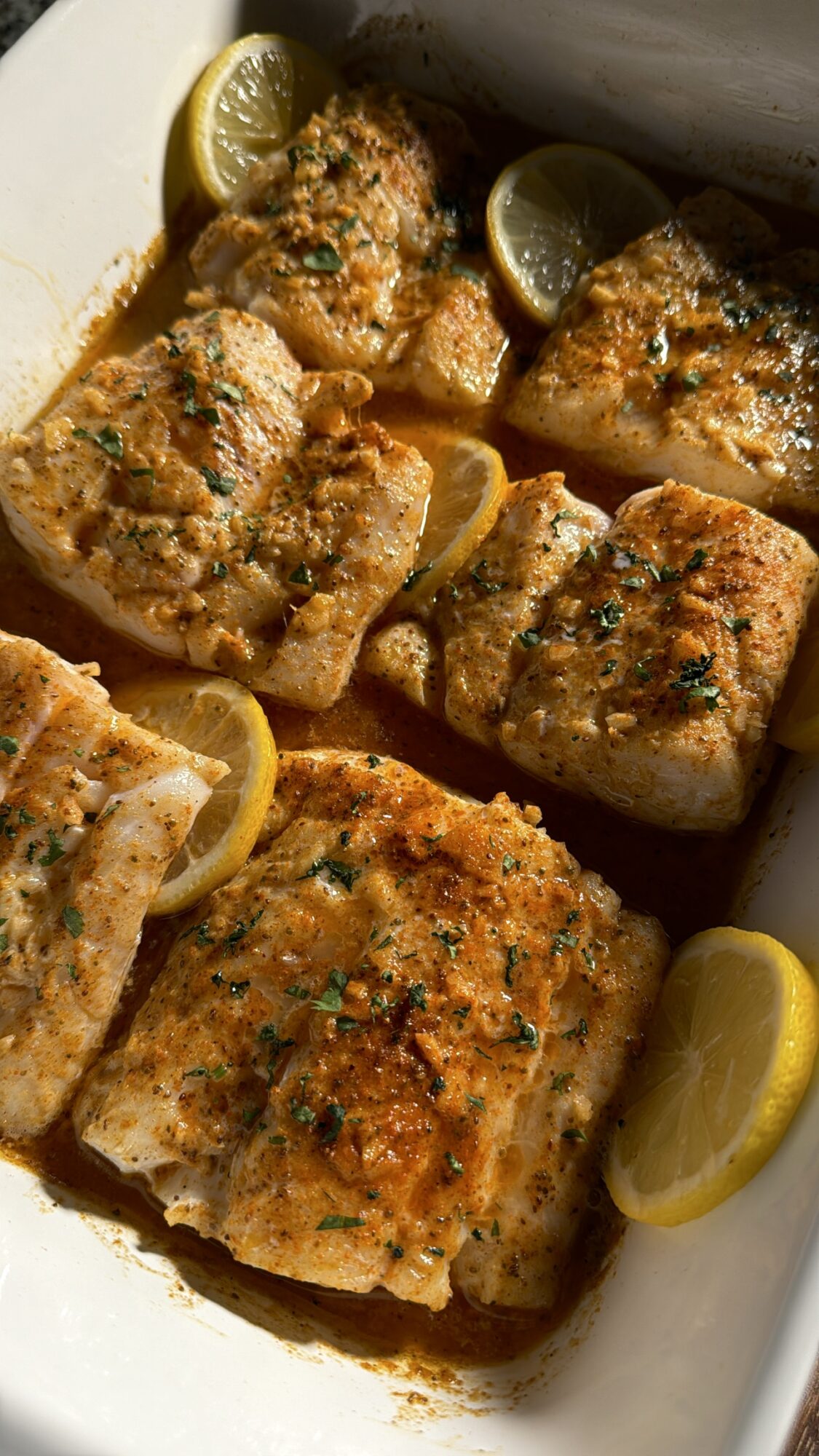 Baked fish fillets garnished with herbs, served with lemon wedges in a white dish.