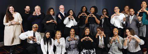 Group of diverse people standing and sitting, smiling, making heart shapes with hands, against a dark background.